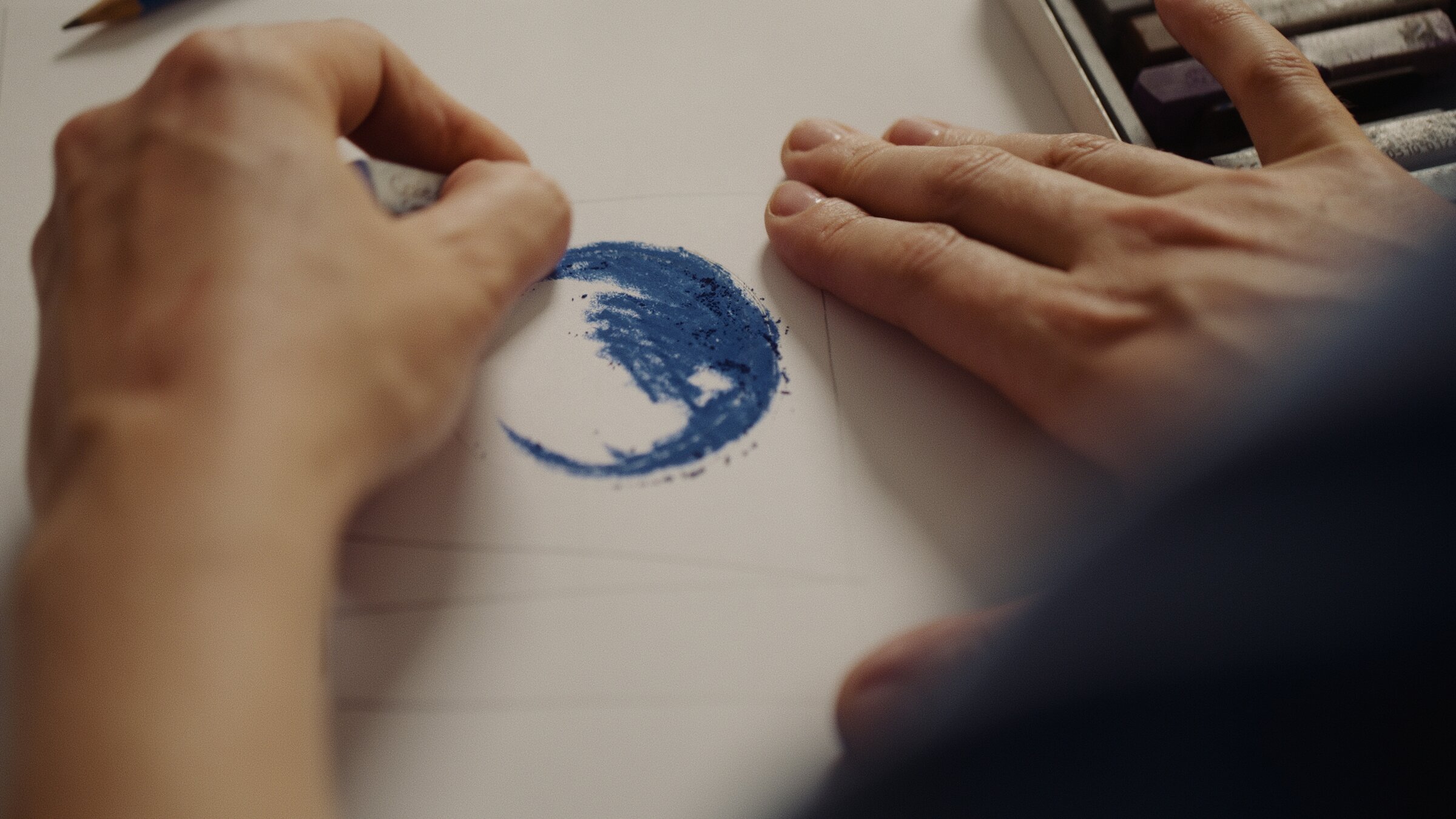 Hands blending a blue pastel circle on paper, palette of pastels alongside