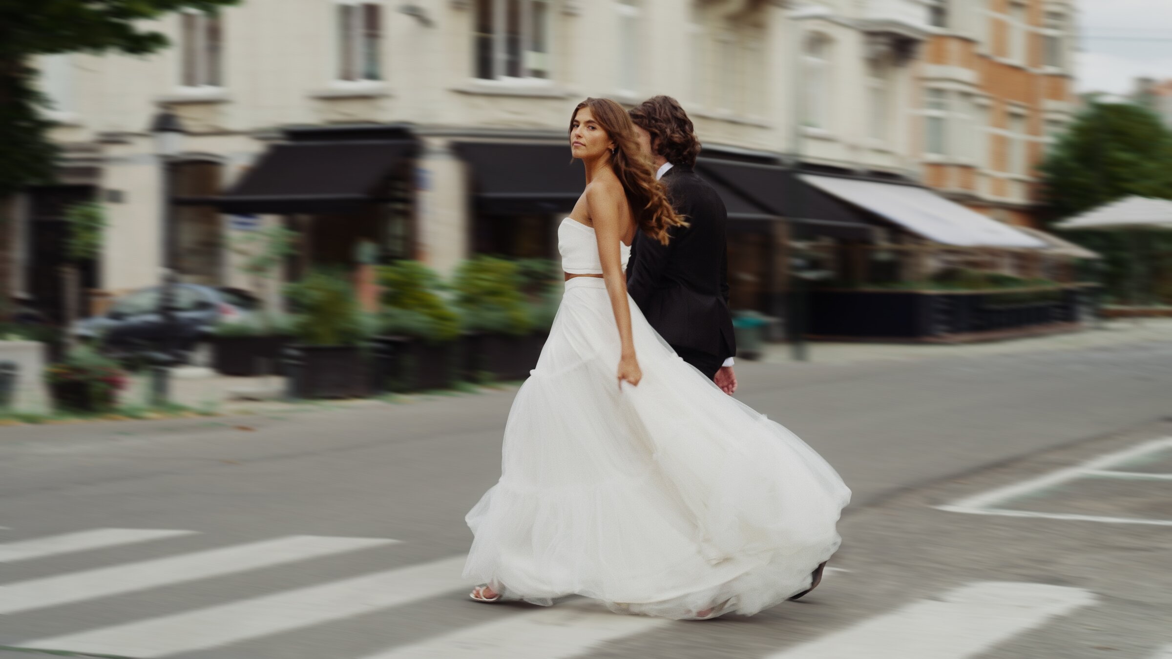 Bride in a strapless gown crossing a city street, groom behind her in motion