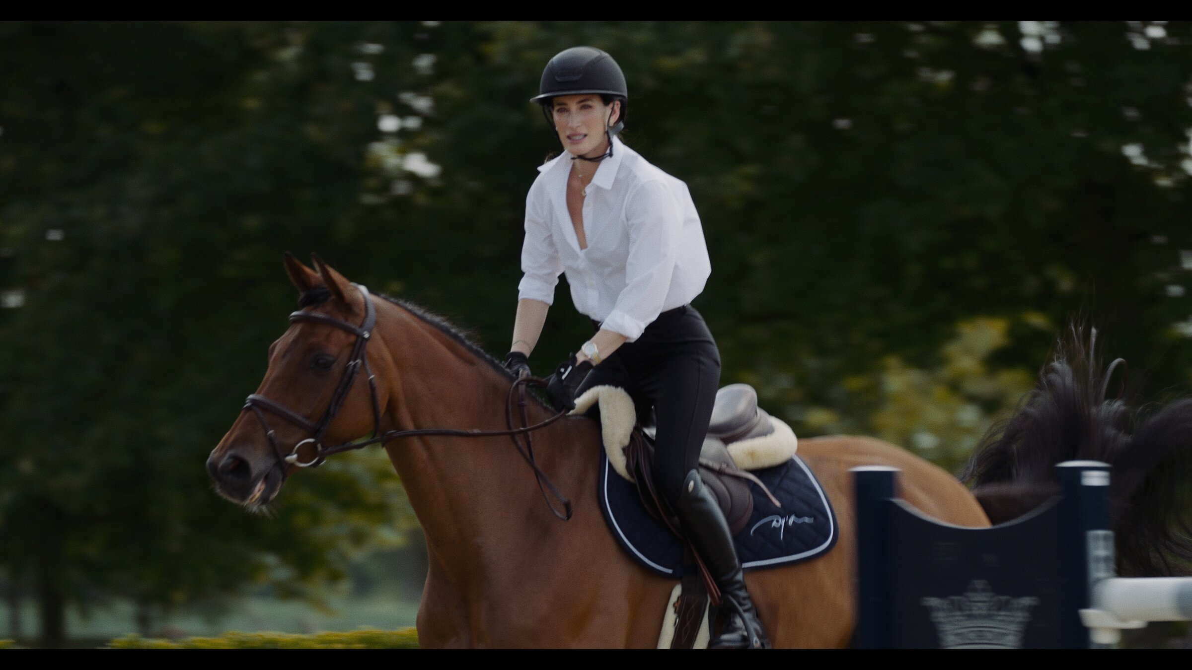 Rider in a white shirt cantering past a jump, tree line behind