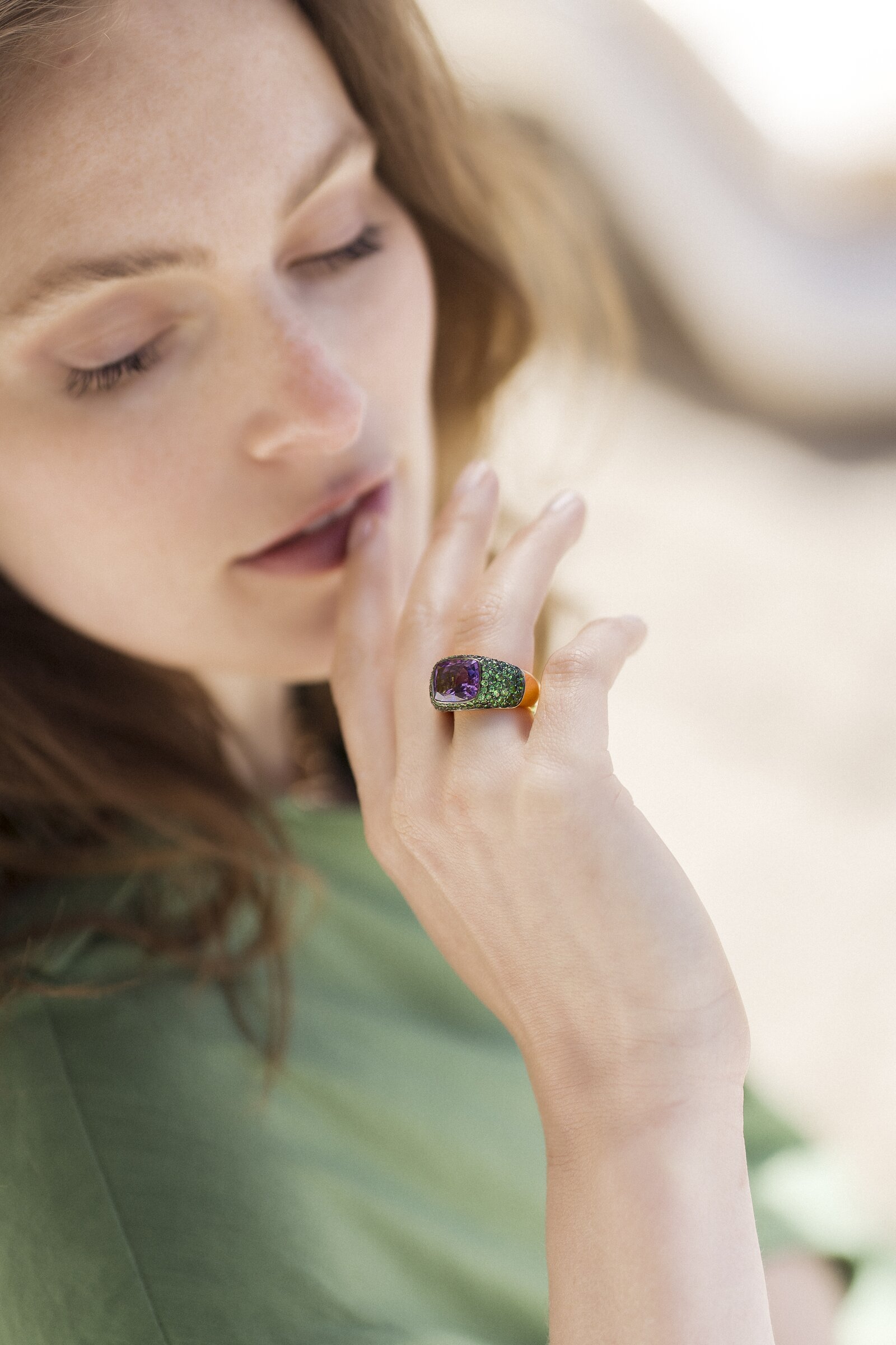 Hand near chin wearing an amethyst and green pavé cushion ring, golden-hour beach light